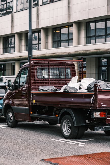 A burgundy flatbed van parked on a city street in front of a modern office building with large glass windows. The van's cargo area is filled with various household items, including wrapped cardboard boxes, furniture covers, and plastic bags, indicating packing materials used for a home relocation. The load is secured with a metal safety barrier mounted at the back of the van. The scene shows the vehicle in the process of either loading or unloading items, illustrating typical furniture transport and moving logistics. Nearby, a white van is partially visible, and a streetlamp is positioned behind the vehicle. The environment appears to be during daylight hours with natural lighting, and the setting suggests a professional removals or moving service such as Man With a Van Teddington, specializing in house removals and packing and moving services.