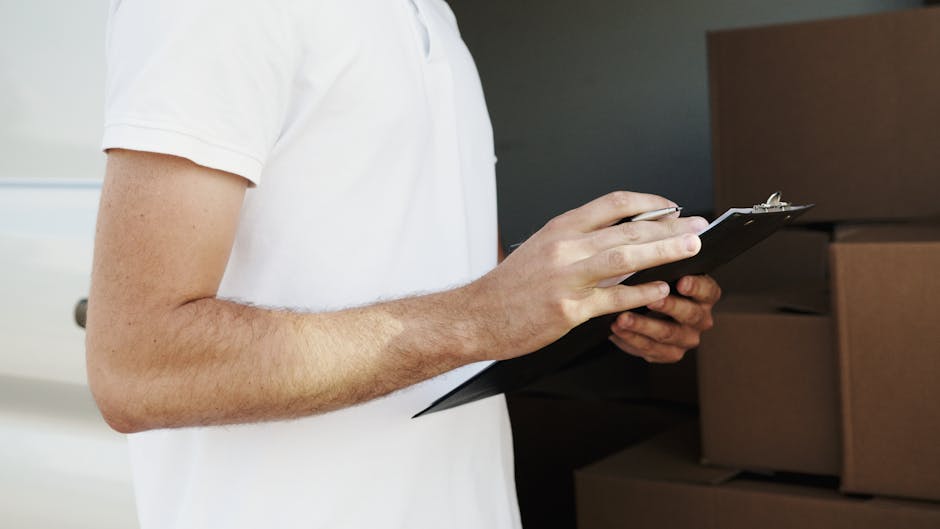 Close-up of a person wearing a white T-shirt, holding and writing on a black clipboard with a pen, standing next to stacked cardboard boxes of various sizes inside a home or storage area. The background includes a white door and a dark wall, suggesting a preparatory stage of home relocation or packing process. The environment is well-lit, with clear visibility of the person's arm and the boxes, reflecting the logistics involved in furniture transport and packing during a house move, as handled by Man With a Van Teddington.