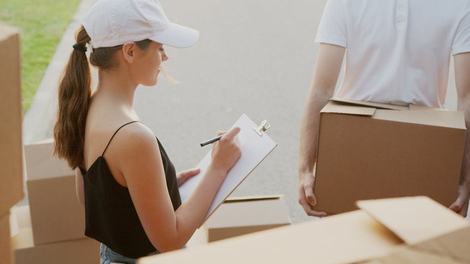 Close-up of a person wearing a white T-shirt, holding and writing on a black clipboard with a pen, standing next to stacked cardboard boxes of various sizes inside a home or storage area. The background includes a white door and a dark wall, suggesting a preparatory stage of home relocation or packing process. The environment is well-lit, with clear visibility of the person's arm and the boxes, reflecting the logistics involved in furniture transport and packing during a house move, as handled by Man With a Van Teddington.