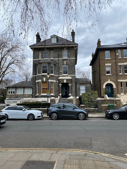 A multi-storey Victorian-style house with a dark brick facade, located on Kingston Road in Teddington, featuring large bay windows on the ground and first floors, and a small central entrance with a black door. The property is positioned along a residential street with several parked cars, including a white vehicle, a dark grey car, and a black vehicle parked directly in front of the house. The street has a curb with a yellow line marking and a tarmac road surface. The surroundings include neighboring houses with similar architectural styles, some with brick facades and bay windows, and a leafless tree with branches extending across the sky. The scene is overcast with grey clouds. Throughout the image, there are no visible moving activities, but the presence of parked vehicles and a residential setting suggest potential ongoing or planned home relocation or furniture transport activities, which [COMPANY_NAME] in Teddington facilitates through its house clearance and moving services.