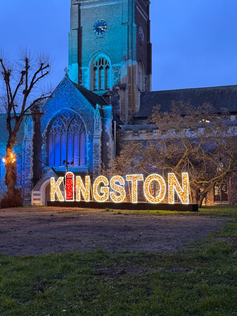 A large illuminated sign spelling 'KINGSTON' in brightly lit, multicoloured letters situated on the grass in front of a historic stone church with tall gothic windows and a clock tower. The church is lit with blue and white lights, and there are leafless trees nearby, indicating a winter evening. The ground appears slightly damp and the sky is dark, suggesting night-time. The scene likely depicts a location serviced by Man With a Van Teddington, which specialises in house removals and furniture transport, with the sign possibly serving as a landmark near the property for a home relocation or house clearance event.