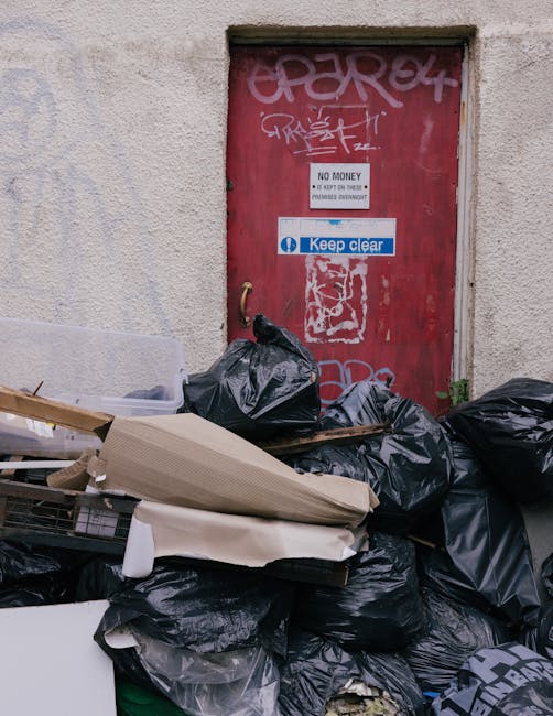A large pile of black rubbish bags containing household waste is stacked against a weathered, red metal door covered with graffiti and various signs, including one that says 'No money' and another instructing to 'Keep clear,' located on the exterior wall of a building. The bags are filled with packaging materials, discarded furniture parts, and other household items, some of which include cardboard boxes, plastic wrap, and pieces of wood. The area appears to be an outdoor space, possibly a back alley or side yard, with concrete paving beneath the waste. This scene relates to the clearance and removal of bulky waste during a home relocation or property clearance service provided by Man With a Van Teddington, which specialises in house removals and transport logistics to manage waste disposal efficiently and in compliance with local regulations.