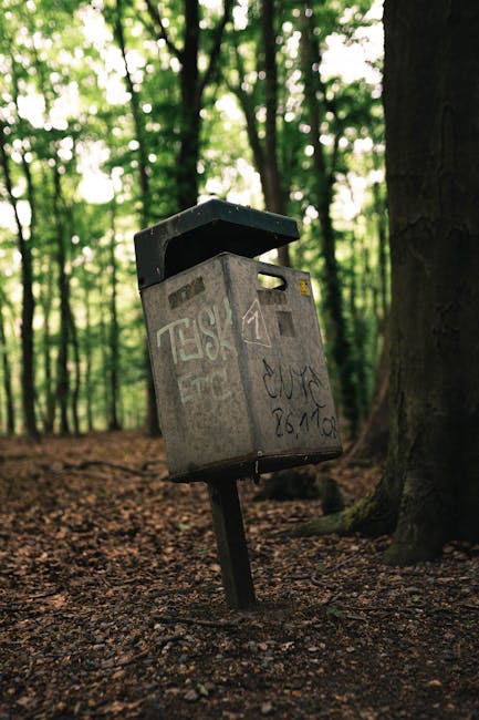 A large pile of black rubbish bags containing household waste is stacked against a weathered, red metal door covered with graffiti and various signs, including one that says 'No money' and another instructing to 'Keep clear,' located on the exterior wall of a building. The bags are filled with packaging materials, discarded furniture parts, and other household items, some of which include cardboard boxes, plastic wrap, and pieces of wood. The area appears to be an outdoor space, possibly a back alley or side yard, with concrete paving beneath the waste. This scene relates to the clearance and removal of bulky waste during a home relocation or property clearance service provided by Man With a Van Teddington, which specialises in house removals and transport logistics to manage waste disposal efficiently and in compliance with local regulations.