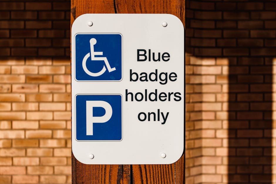 Close-up image of a rectangular white parking sign mounted on a wooden post with four screws, situated in front of a red brick wall. The sign features two sections: the top section displays a blue background with a white wheelchair symbol, indicating accessible parking, and the bottom section has a large white letter 'P' on a blue background, signifying parking area. To the right of the wheelchair symbol, the text reads 'Blue badge holders only,' indicating that parking is restricted for authorized permit holders. The sign is outdoors, and sunlight casts shadows on the brick wall background. This parking sign pertains to regulations related to house removals and the transportation of furniture, supporting moving logistics and home relocation services provided by companies like Man With a Van Teddington, especially in contexts where parking permits or restricted access areas are relevant for effective house removals.