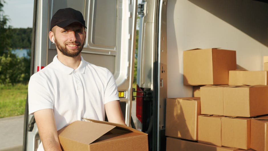 A man wearing a white t-shirt, blue jeans, and a black cap is sitting on the pavement beside an open van, which is loaded with numerous cardboard boxes of various sizes, some sealed with packing tape.
