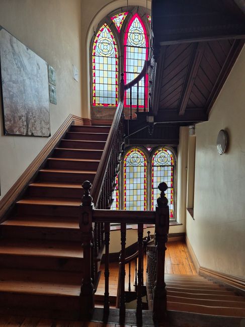 Interior view of a historic Victorian home staircase during a home relocation process, featuring dark wooden stairs with a polished handrail and turned balusters. The staircase is positioned adjacent to large, colorful stained-glass windows with intricate patterns, allowing natural light to illuminate the area. A portion of the staircase continues upward, with a landing visible behind the stained glass. Inside the hallway, there are light-colored walls with minimal decoration, including a wall-mounted light fixture and a painting on the wall. The setting appears to be inside a residence, with a clean and well-maintained environment, reflecting the type of home where professional removals services like those provided by Man With a Van Teddington are often utilized for furniture transport and moving logistics, especially in buildings with traditional architecture requiring careful navigation around tight spots and delicate features.