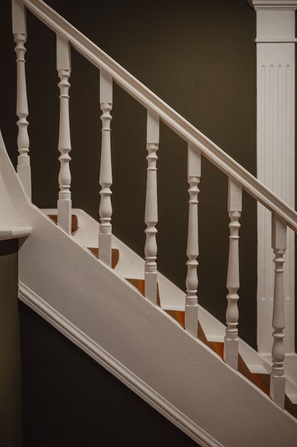 A close-up view of a staircase in a Victorian home interior, featuring white wooden balustrades and handrails, with visible wooden steps finished in a natural brown tone. The staircase curves upward, occupying a central position in the image, with a dark green wall in the background and soft, natural lighting illuminating the woodwork and creating subtle shadows. The surrounding area includes parts of a white wooden newel post and adjacent wall, with no visible moving equipment or packaging materials present. This setting exemplifies a typical home interior where professional removals, such as those provided by Man With a Van Teddington, might carefully navigate tight-spots during a house relocation, ensuring furniture transport and packing are performed with precision within confined stairwell spaces.