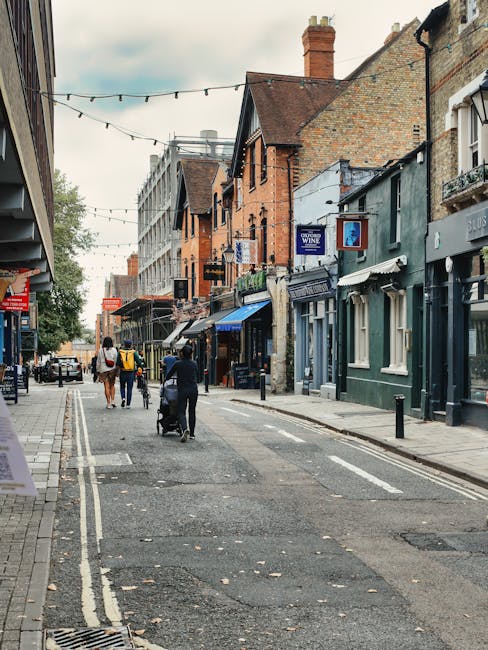 A narrow street lined with mixed-style terraced and commercial buildings, featuring brick and painted facades, with several storefronts including a wine shop and a café, all above a pavement with pedestrians walking and pushing strollers. A man from Man With a Van Teddington is seen loading or unloading moving boxes and furniture wrapped in cardboard and plastic onto a vehicle parked at the curb, utilizing trolleys and blankets for protection. The scene captures the typical environment of a home relocation or furniture transport process, with streetlights and overhead string lights enhancing the daytime setting. The surroundings reflect a busy urban area suitable for house removals and transport services operating in tight urban access conditions, highlighting the importance of careful planning for furniture transport through narrow streets.