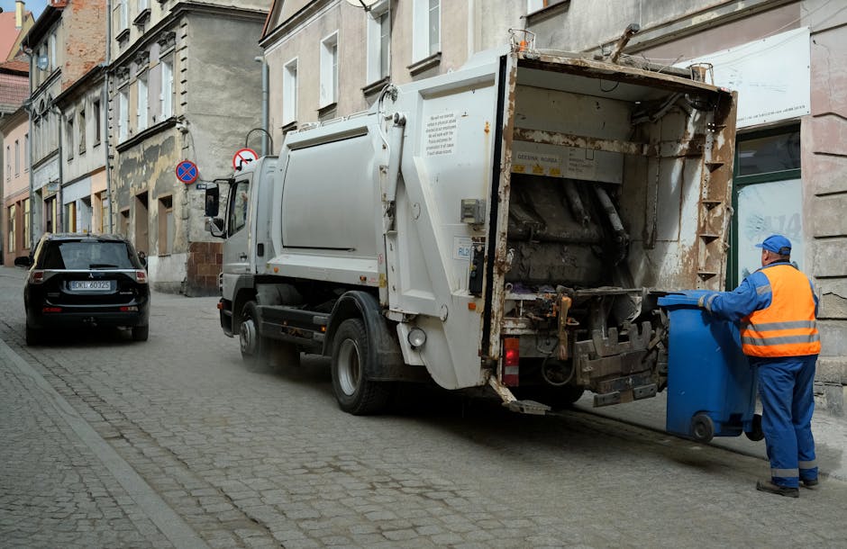 A street scene showing a refuse collection truck from Man With a Van Teddington positioned parallel to the pavement outside a row of residential buildings with traditional brick facades. The rear of t