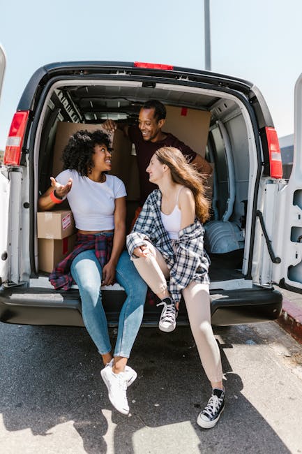 Inside the open cargo area of a large, modern moving van, two young women sit on the edge of the loading floor engaged in conversation, while a man stands behind them, smiling. The interior of the van contains several cardboard boxes and wrapped items prepared for moving, with some visible clothing and blankets being used for padding. The van is parked outdoors on a paved surface, with the bright sky overhead, indicating daytime. Metal rails and straps are seen securing the load, and the van’s rear doors are wide open, revealing the interior space designated for furniture transport during a home relocation. The scene captures the final stages of packing and sorting items for a house removal, reflecting a professional and organized moving process that Man With a Van Teddington might facilitate for clients planning to move from a TW11 flat or apartment, emphasizing the packing, loading, and transportation phases of removals.
