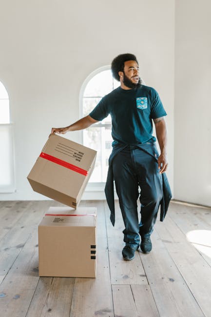 A man with dark skin and a beard, wearing a blue T-shirt with a logo and dark trousers, is inside a bright, minimally furnished room with white walls and a large arched window. He is holding a cardboard box decorated with red tape, preparing to lift or place it on top of another box on the wooden floor. Nearby, there are additional packed boxes, some wrapped in plastic or packing paper, indicating an ongoing house relocation process. The natural light from the window highlights the clean, uncluttered environment, and the scene reflects furniture transport and packing activities associated with professional removals, as offered by Man With a Van Teddington, focusing on efficient household moves and logistical planning for flat relocations in Tw11.
