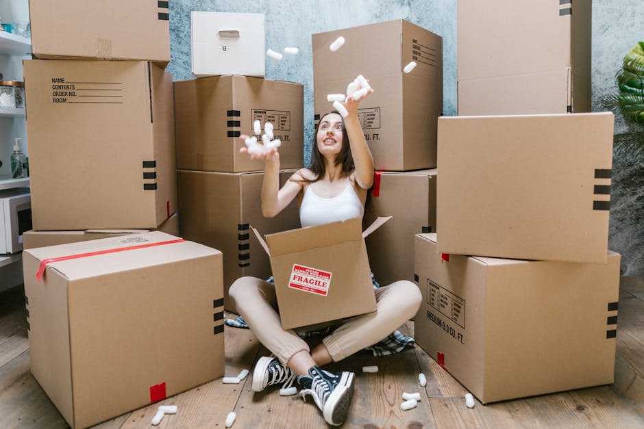 Inside the open cargo area of a large, modern moving van, two young women sit on the edge of the loading floor engaged in conversation, while a man stands behind them, smiling. The interior of the van contains several cardboard boxes and wrapped items prepared for moving, with some visible clothing and blankets being used for padding. The van is parked outdoors on a paved surface, with the bright sky overhead, indicating daytime. Metal rails and straps are seen securing the load, and the van’s rear doors are wide open, revealing the interior space designated for furniture transport during a home relocation. The scene captures the final stages of packing and sorting items for a house removal, reflecting a professional and organized moving process that Man With a Van Teddington might facilitate for clients planning to move from a TW11 flat or apartment, emphasizing the packing, loading, and transportation phases of removals.