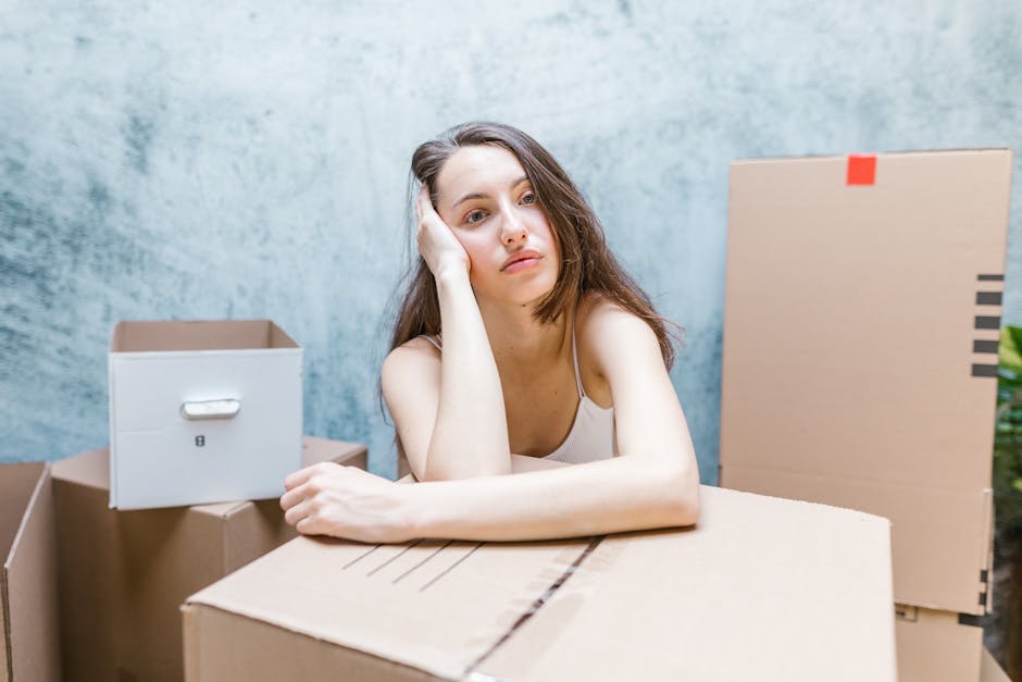 A young woman with long brown hair and a neutral expression rests her head on her left hand while leaning on a large cardboard box in a room filled with packed moving boxes. The boxes are made of brown and white cardboard, some sealed with packing tape or secured with orange tape, and vary in size. The room features a light blue textured wall and natural lighting. Inside the space, there is a doorway visible on the right, leading to an outdoor area. The scene depicts the preparation and packing process associated with house removals, as part of a home relocation or furniture transport service conducted by Man With a Van Teddington. The woman appears to be waiting during the packing or loading activity typical of a moving process involving careful handling of belongings before transport.