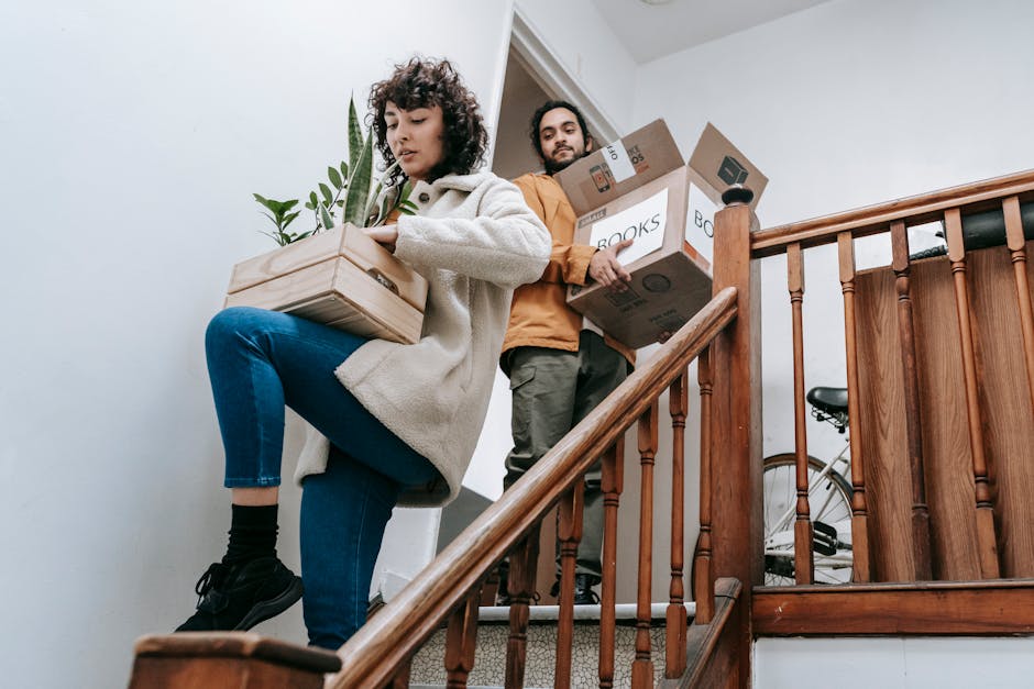 A woman with curly dark hair, dressed in a cream-colored fleece jacket, blue jeans, and black shoes, is carrying a small cardboard box with a potted plant on top, ascending a wooden staircase inside a home. Behind her, a man with dark hair and a beard, wearing a brown jacket and dark pants, is holding three medium-sized cardboard boxes labeled 'BOOKS' and 'FRAGILE,' preparing to move them up the stairs. The staircase features a polished wooden handrail and spindles, with a bicycle partially visible behind the man on the landing. The space is well-lit with natural light, and the walls are painted white. The scene depicts a professional home relocation process managed by Man With a Van Teddington, involving packing, lifting, and transporting household belongings during an internal move or home relocation, with attention to safe handling and organized moving logistics.