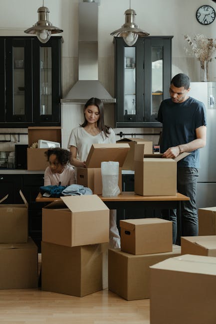 A young woman with long brown hair and a neutral expression rests her head on her left hand while leaning on a large cardboard box in a room filled with packed moving boxes. The boxes are made of brown and white cardboard, some sealed with packing tape or secured with orange tape, and vary in size. The room features a light blue textured wall and natural lighting. Inside the space, there is a doorway visible on the right, leading to an outdoor area. The scene depicts the preparation and packing process associated with house removals, as part of a home relocation or furniture transport service conducted by Man With a Van Teddington. The woman appears to be waiting during the packing or loading activity typical of a moving process involving careful handling of belongings before transport.